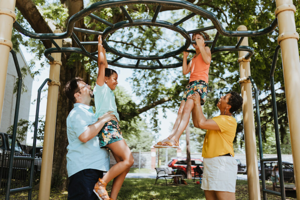 Two parents are helping their kids play on the monkey bars at the park